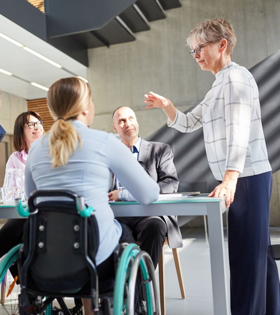 1 standing woman talking to 3 persons sitting on a table